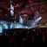 A large indoor event with a stage featuring bright screens and spotlights, attended by a substantial crowd. The venue has a high ceiling with industrial beams and red lighting accents. The atmosphere resembles a concert or a conference with a focus on visual effects.